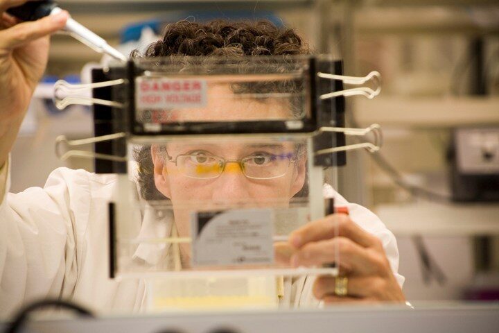 Dr. Christopher Pearson in the laboratory wearing a white lab coat and protective glasses carefully handles a glass container secured with multiple black binder clips.