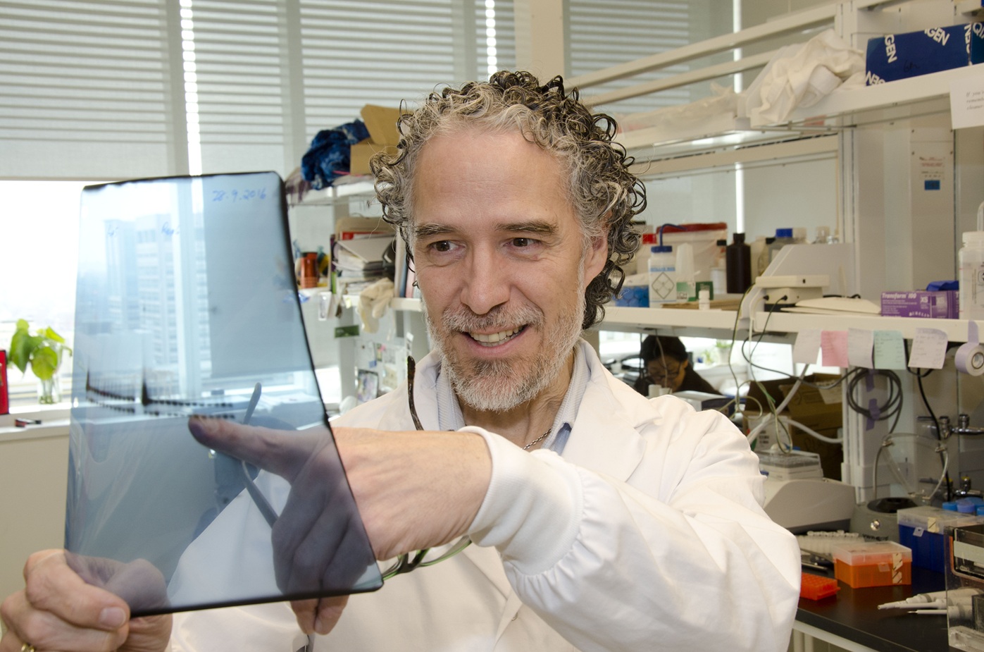 Dr. Christopher Pearson in a white lab coat, smiling as he points to a translucent blue autoradiograph film in a research lab setting