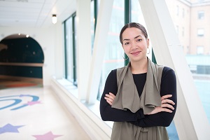 Woman wearing black long sleeve shirt and gray cardigan, standing and smiling with her arms crossed in a hallway. 