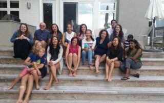 A group of people sit on some steps in front of a cottage, smiling at the camera.