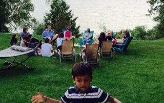 A young boy stands in front of a lakeside gathering, where people are seated on lawn chairs facing the water.