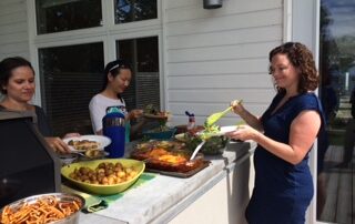 A group of women serving themselves food on a counter outdoors, next to a cottage.