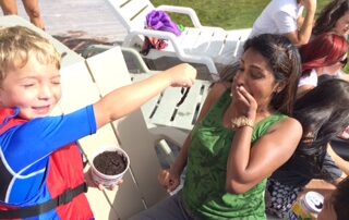 smiling boy holds up a worm near a surprised woman on a sunny deck.