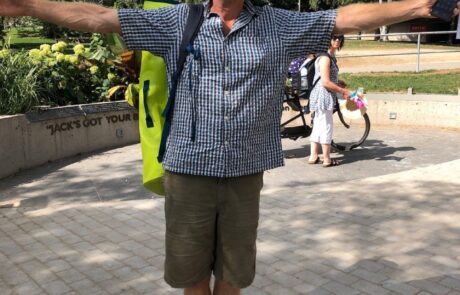 Man in a sun hat, sunglasses, and checkered shirt standing outdoors with arms outstretched, smiling. He has a green neon backpack over one shoulder.