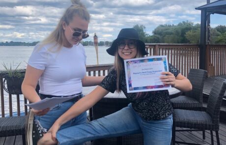 Two people are outdoors on a deck by the water. One person is holding a colorful certificate and smiling, wearing a black hat and sunglasses, while playfully lifting a leg. The other person, in a white shirt and sunglasses, is holding papers and looking down