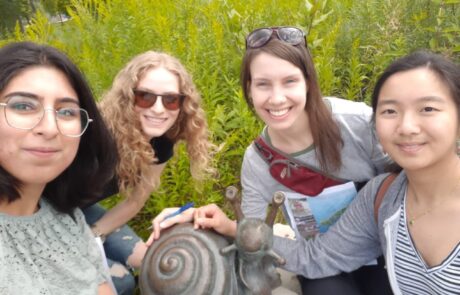 The image shows four people smiling at the camera while posing next to a decorative snail sculpture. They appear to be outdoors, surrounded by green plants.