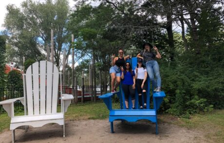 five people standing on a very large blue Adirondack-style chair. There’s another oversized white chair beside it. The setting appears to be outdoors in a park or recreational area with trees and a fence in the background.