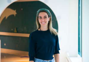 Woman with brunette hair and a headband, wearing a dark blue sweater, standing in a hallway, smiling with her hands by her side.