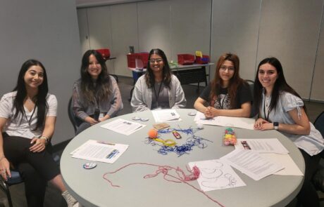 Group of 5 girls sitting at a round table smiling with paper and string on the table.