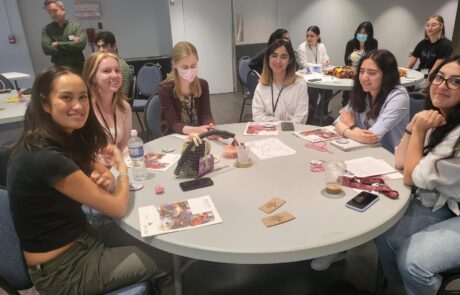 Group of 6 girls sitting at a round table smiling with paper and string on the table.
