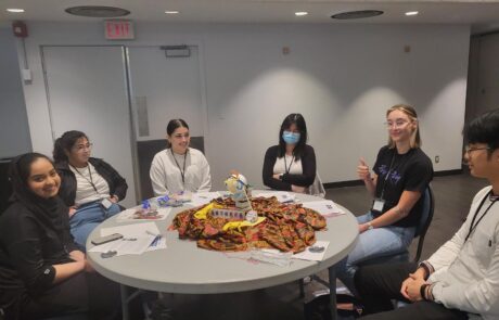 Group of 6 girls sitting at a round table smiling with paper and string on the table.