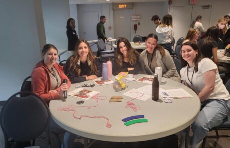 Group of 5 girls sitting at a round table smiling with paper and string on the table.
