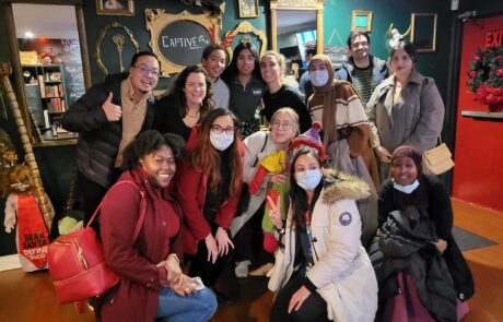 A group of people posing for a photo inside a venue with a green wall and framed decor. Some are wearing winter jackets, and a few are wearing masks. A chalkboard sign in the background reads 'CAPTIVE