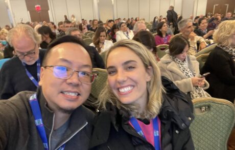 Two attendees smiling for a selfie at a crowded conference session, wearing lanyards. Rows of seated participants fill the background, some engaged with the event and others using their phones.