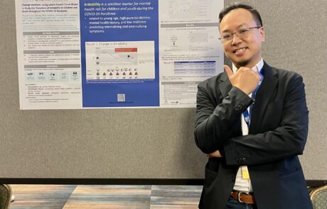 Researcher smiling in front of a scientific research poster, wearing name badges and lanyards, in a professional conference setting