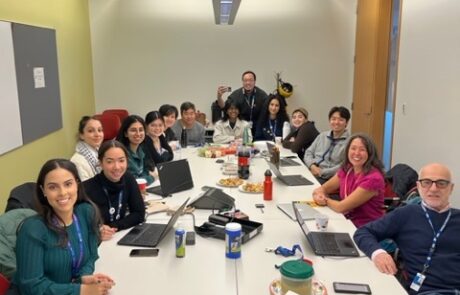 Group of people sitting a large desk in a large conference room with laptops in front of them, smiling.