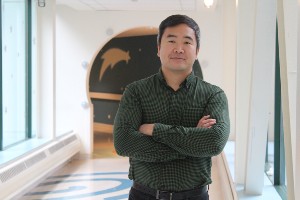 Alex standing in a hallway, front-facing, smiling with his arms crossed.