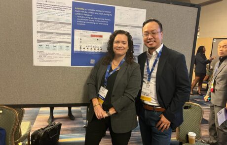 Two conference attendees smiling in front of a scientific research poster, wearing name badges and lanyards, in a professional conference setting.