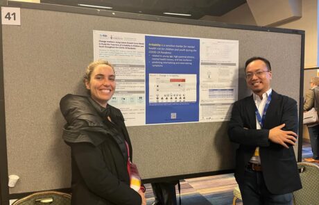 Two conference attendees smiling in front of a scientific research poster, wearing name badges and lanyards, in a professional conference setting