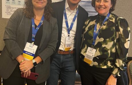 Three researchers standing in front of a poster, wearing lanyards and smiling at the camera.
