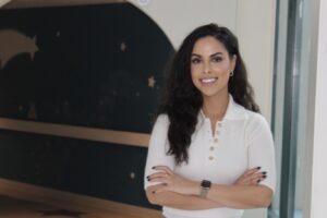 Woman with curly dark hair and a white floral‑embroidered cardigan, standing and smiling in front of a circular archway painted with a shooting star motif.