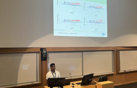 Man with white shirt and lanyard behind a podium presenting a powerpoint projected behind him in a lecture hall.