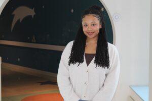Woman with long black locs, wearing a cream-colored cardigan over a brown top, stands smiling in front of a circular archway with star and shooting star wall art.