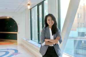 Woman with straight black hair wearing a white shirt and gray blazer, standing in a hallway, smiling with her arms crossed. 