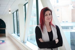 Woman with red dyed hair wearing a black shirt and white cardigan, standing and smiling in a hallway.
