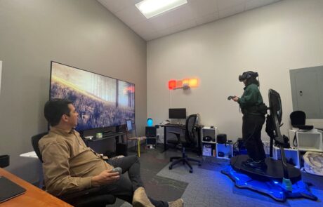 A woman wearing a virtual reality headset and holding controls, standing on a virtual reality platform while a man sits on a chair and watches.
