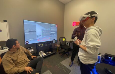 One man wearing a virtual reality headset and holding virtual reality controls. Two other men on either side of him and a TV, one is sitting, the other is standing.