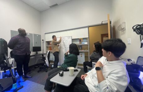 Three people sitting down in chairs in a room, looking at a man point to a white board beside him. Another man is also standing on the left.