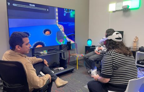 A woman sitting, wearing a virtual reality headset and holding controls facing a TV. Two people on either side are also sitting, watching her.