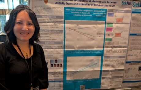 Woman with short black hair, smiling and standing beside a research poster titled "Facial emotion recognition moderates link between autistic traits and irritability in children and youth".