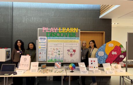 A table titled "play learn discover" with various posters, infographics, and games. Two woman stand on either side of the booth.