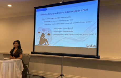 A woman sits at a table beside a projector screen with a powerpoint presentation projected on.
