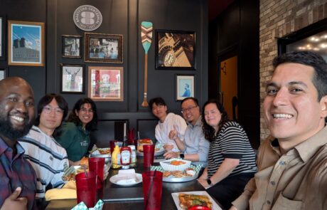 7 people take a selfie at a restaurant dinner table. There is food and drinks on the table and they are all looking at the camera and smiling.