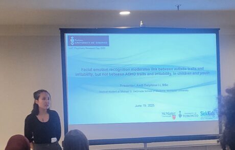 A girl in a black long sleeve shirt stands beside a project screen presenting a powerpoint, she is looking off camera.