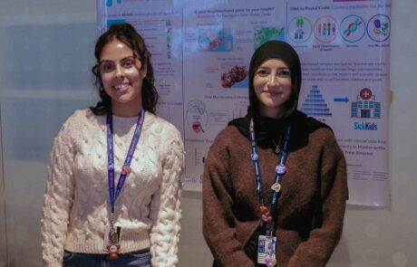 Two women wearing lanyards, stand in front of a research poster, smiling at the camera.