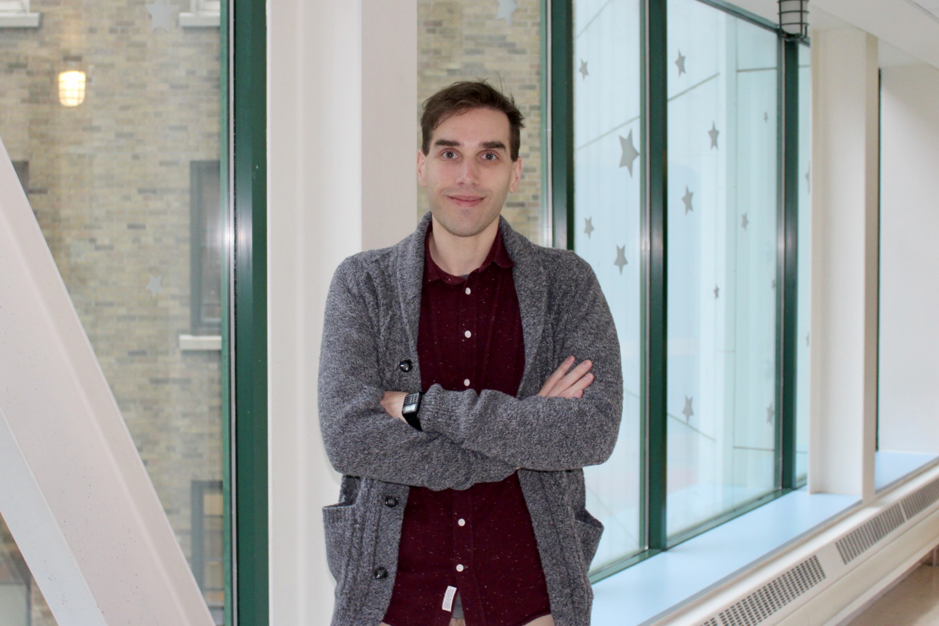 Man with red button up shirt and gray cardigan standing in front of windows smiling at the camera, with his arms crossed.