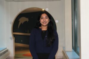Woman with long black locs, wearing a cream-colored cardigan over a brown top, stands smiling in front of a circular archway with star and shooting star wall art.