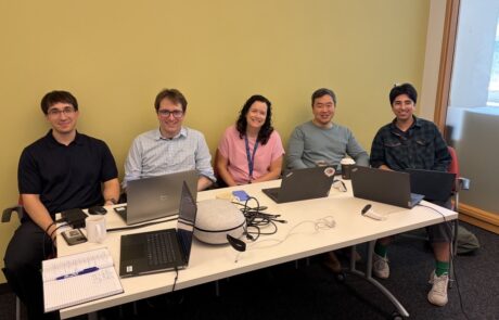5 people sitting at a white table with computers, looking up at the camera and smiling.