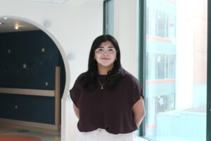 Girll with black hair, burgundy shirt, and white pants standing in front of an archway and window, smiling at the camera.