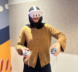 A person wearing a white virtual‑reality headset stands indoors holding two VR controllers.