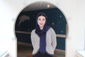 Girl with blue headscarf standing in front of an archway with a striped shirt, smiling at the camera.