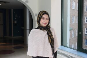 Girl with white poncho top and a black headscarf standing in a hallway, smiling at the camera.