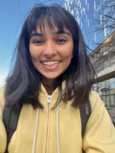 Selfie of a girl with dark brown short hair and bangs wearing a yellow zip up hoodie, smiling at the camera.