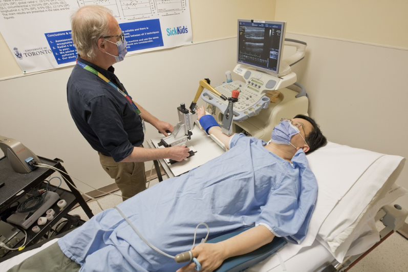 In the vascular ultrasound area the sonographer is operating equipment to acquire images of blood vessels in the patient's arm. The patient is lying comfortably on their back with additional sensors attached to their fingers.