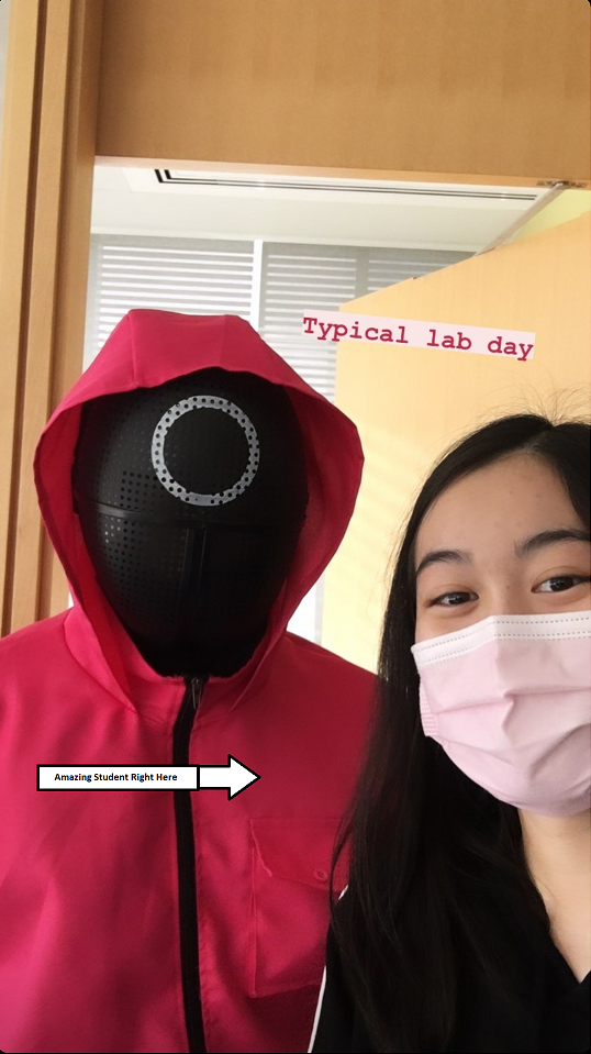 A woman in a mask taking a selfie with a person dressed in a Squid Game costume. The text reads: Typical lab day. There is an arrow pointing toward the woman that reads: Amazing student right here.
