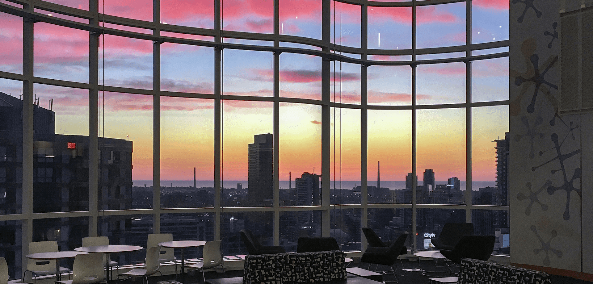 A landscape photograph of the sun rising above buildings in Downtown Toronto, taken from an interactive space in the Peter Gilgan Centre
